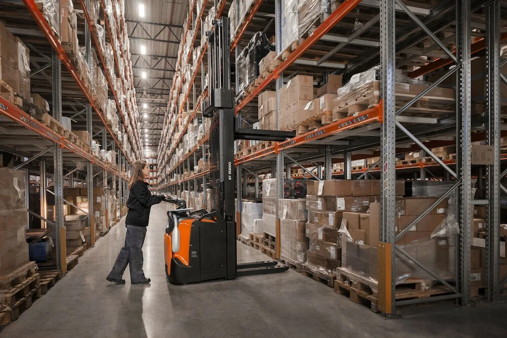 BT Staxio SPE160, electric stacker with platform, and side guards. Application photo in warehouse environment.