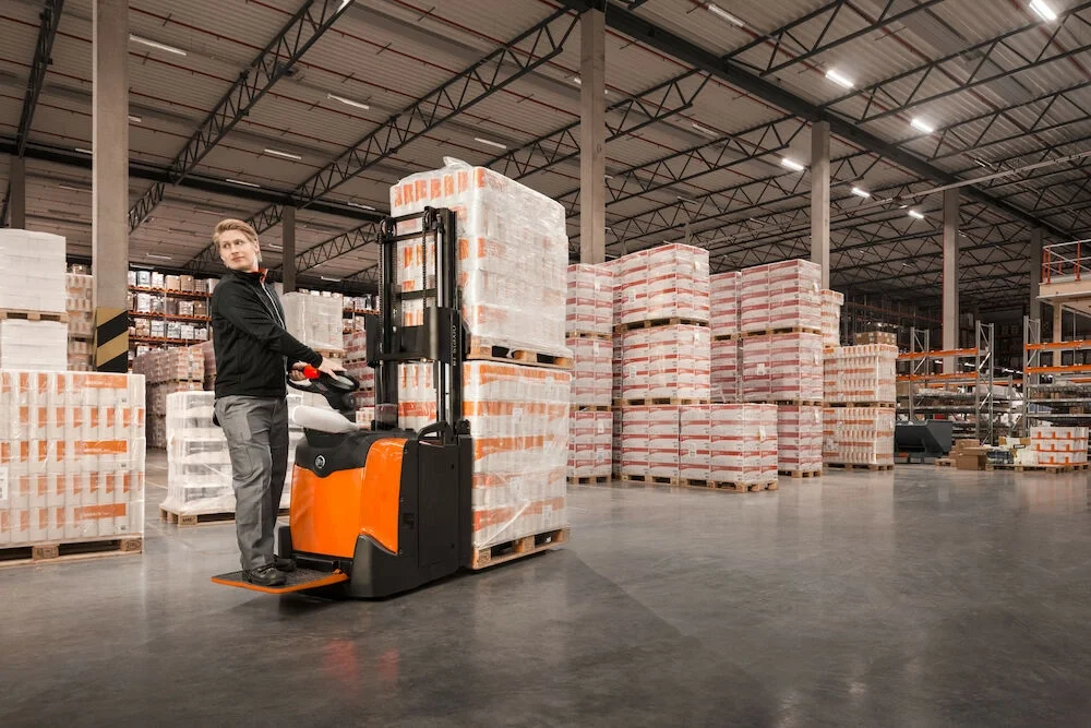 BT Staxio SPE200D, electric stacker with double load handling, platform, and side guards. Application photo in warehouse environment.