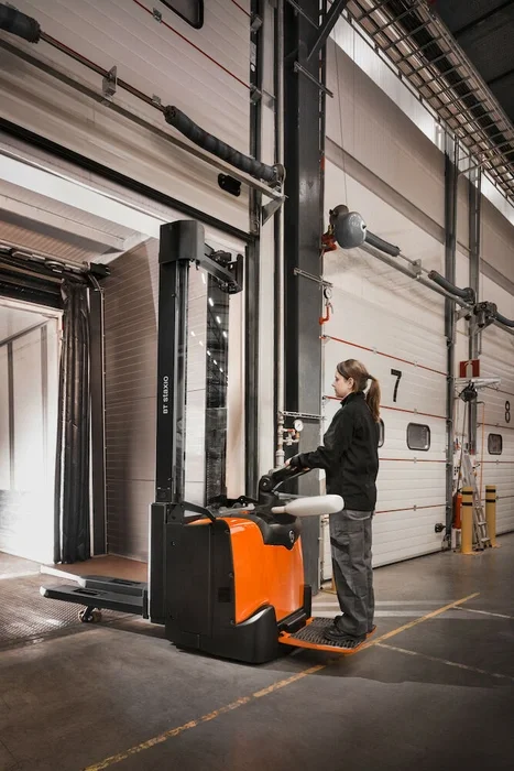 BT Staxio SPE160L, electric stacker with elevating support arms, platform, and side guards. Application photo in warehouse environment.