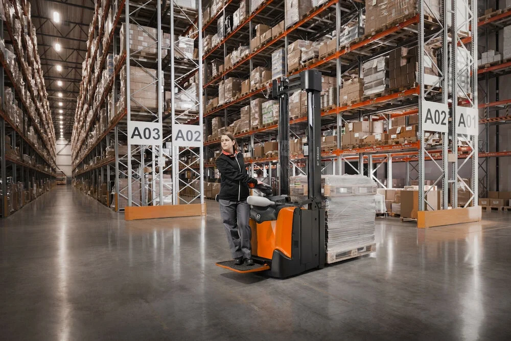 BT Staxio SPE160, electric stacker with platform and side guards. Application photo in warehouse environment.