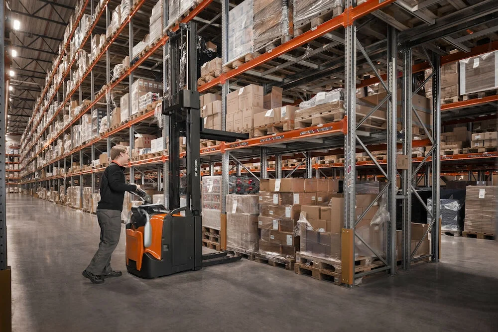 BT Staxio SPE160, electric stacker with platform, and side guards. Application photo in warehouse environment.