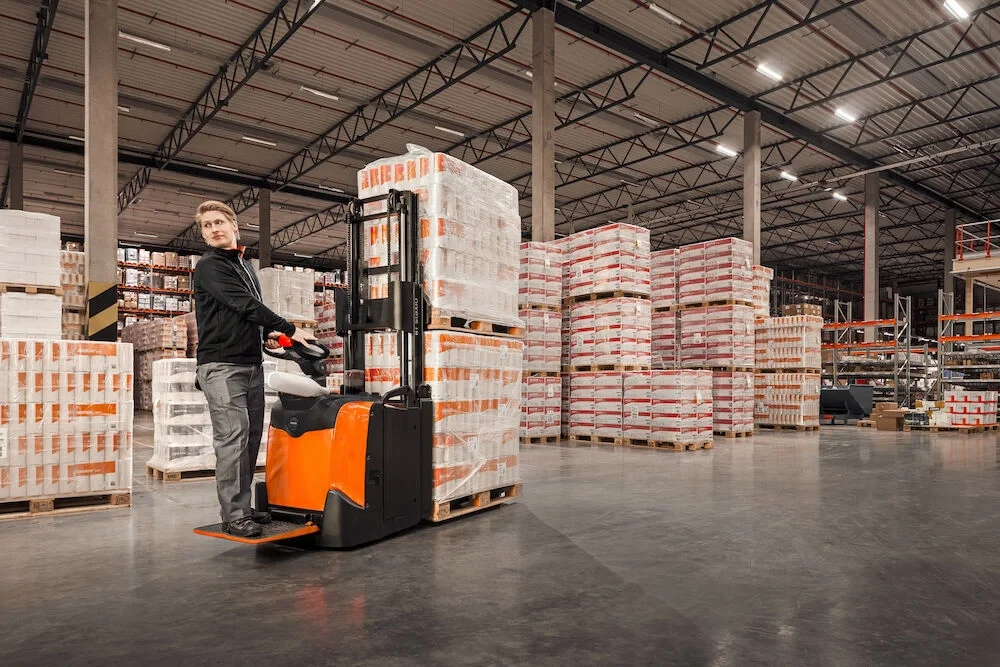 BT Staxio SPE160L, electric stacker with elevating support arms, platform, and side guards. Application photo in warehouse environment.
